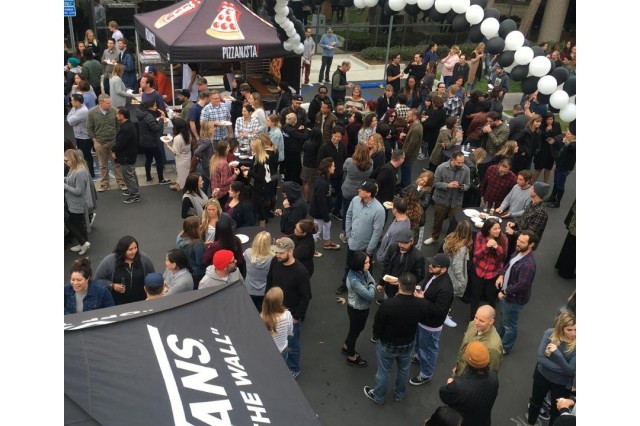 A large crowd of people at an outdoor event with a black and white balloon arch between a Pizzanista1-branded pop-up tent and a Vans-branded pop-up tent.